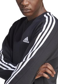 Sweatshirt atlético preto com o logótipo branco da Adidas e três riscas brancas ao longo dos ombros e das mangas. Manga longa com punhos canelados.