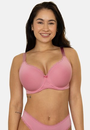 Smiling woman with long dark hair wearing a pink lace-trimmed bra and matching underwear against a plain white background.