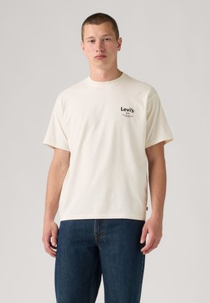 Νέος άνδρας που φοράει απλό off-white μπλουζάκι Levi's και σκούρο μπλε τζιν μπροστά σε ένα απλό ανοιχτό φόντο.