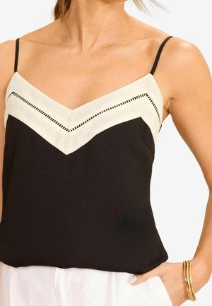 Vrouw die een zwart-wit mouwloos topje met dunne bandjes draagt, gecombineerd met een witte broek en gouden armbanden om haar pols.