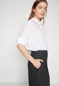Blusa bianca con bottoni e maniche ripiegate, realizzata in tessuto leggero. Abbinata a pantaloni scuri a vita alta con pieghe e tasche laterali.