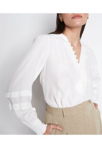 Kvinna som bär en vit blus med knäppt V-halsspatel och lager av puffärmar, kombinerad med hög midja beige byxor, händerna i fickorna.