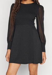 Schwarzes Kleid mit einem gerippten Oberteil und durchsichtigen Puffärmeln. Es hat einen runden Ausschnitt und einen geraden Schnitt, mit einer weichen Textur und minimaler Verzierung.