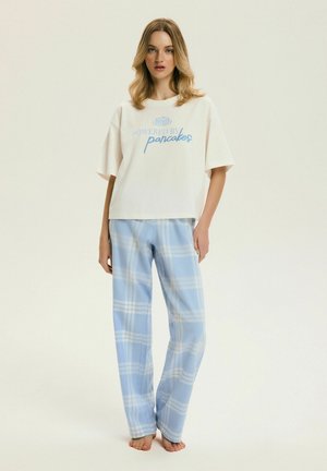 Młoda kobieta stojąca boso, ubrana w białą, oversize'ową koszulkę z napisem „Powered by pancakes” oraz jasnoniebieskie, w kratę spodnie piżamowe.