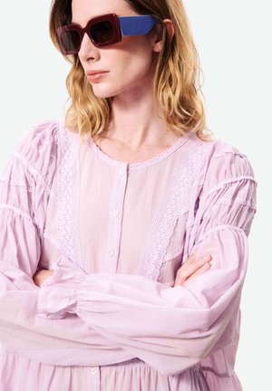 Femme aux cheveux blonds ondulés portant de grandes lunettes rectangulaires aux branches bleues et un chemisier rose pâle orné de dentelle, les bras croisés.