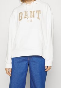 Sudadera blanca con el logo de GANT en beige, capucha con cordón, bolsillo frontal tipo canguro y puños de canalé. Combinada con jeans de mezclilla azul.