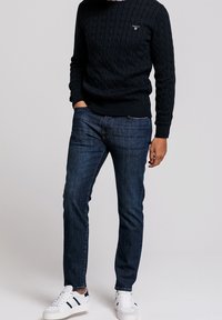 Homme portant un pull à torsades bleu foncé, un jean bleu, des baskets blanches avec des bandes noires, debout avec une main dans la poche sur un fond uni.