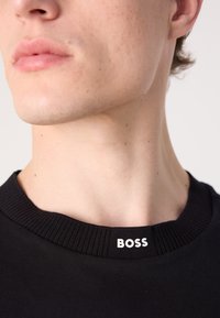 Črni majica s kratkimi rokavi z rebrastim ovratnikom in belim oznako "BOSS" na ovratniku. Pouzdan posnetek detajlov ovratnika in majice.