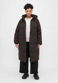 Brown long puffer coat with a hood, featuring a quilted design, zipper closure, and side pockets, worn over a light sweater and wide pants.