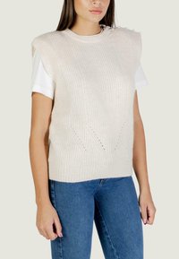 Gilet in maglia di un beige chiaro con una texture a coste, dotato di bottoni sulle spalle e una vestibilità rilassata, indossato sopra una camicia bianca.