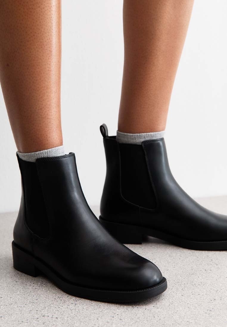 Gros plan de jambes portant des bottines en cuir noir avec des chaussettes grises sur un sol clair et un fond uni.