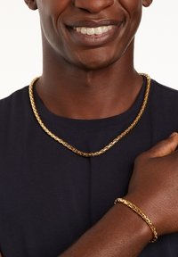 Gouden ketting en armband, beide met een gestructureerd ontwerp met schakels. De ketting ligt op een donker shirt, wat hun glans benadrukt.