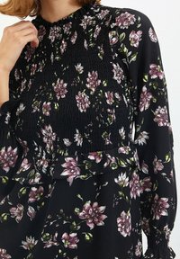 Kobieta ubrana w czarną sukienkę w kwiaty z różowymi i białymi kwiatami, z marszczonym dekoltem oraz długimi rękawami z elastycznymi mankietami.