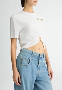 T-shirt blanc à manches courtes avec un centre froncé et un détail de logo doré, associé à un jean en denim bleu clair taille haute.