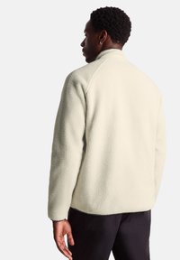 Fleece jacket in off-white with a textured surface, high collar, and raglan sleeves, worn over dark pants. Simple, functional design.
