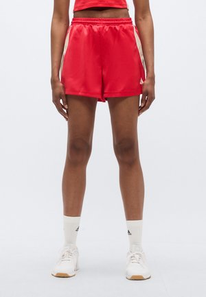 Person iført røde atletikshorts, hvide sokker med sort logo og hvide sneakers, stående mod en ensfarvet baggrund.