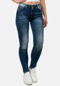 Jeans in denim skinny scuro con dettagli sbiaditi. Presentano una chiusura anteriore con bottone e tasche frontali. Abbinati a sneakers bianche.