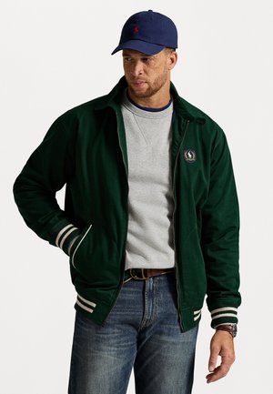Man in een marineblauwe pet, een donkergroene ritsjack met gestreepte manchetten, een lichtgrijze sweatshirt, een blauwe jeans en een bruine horloge op een witte achtergrond.