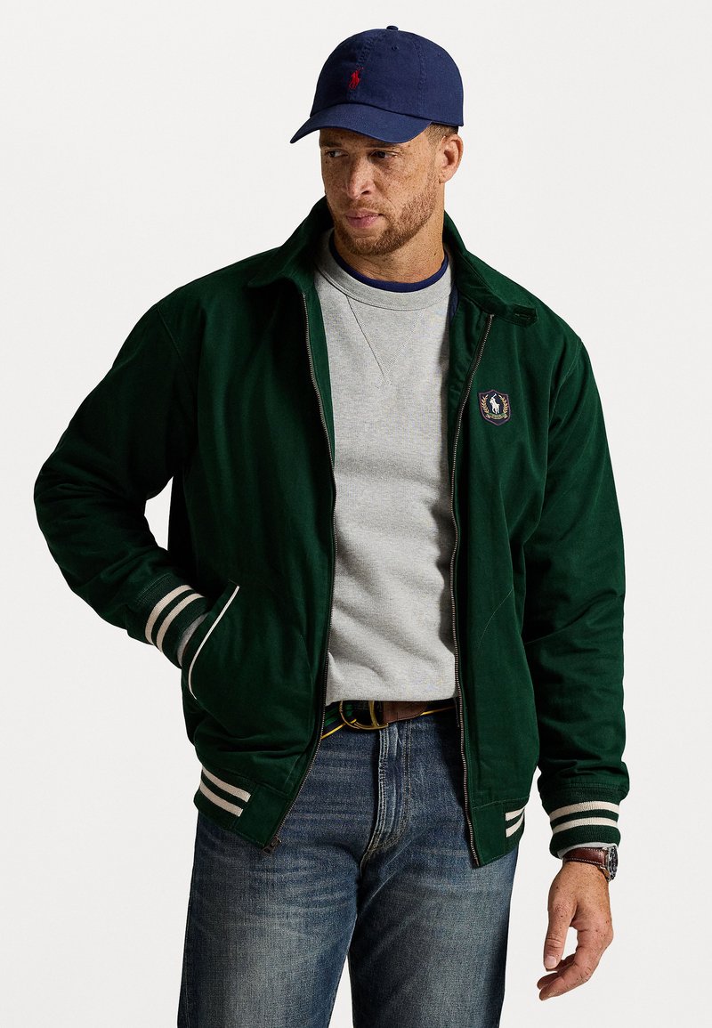 Mand iført en marineblå kasket, mørkegrøn zip-up jakke med stribede manchetter, lysegrå sweatshirt, blå jeans og brunt ur på hvid baggrund.