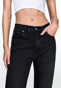 Jeans de mezclilla negra de cintura alta, diseño de cinco bolsillos y acentos de hardware de cobre. La textura es suave con un ligero brillo.