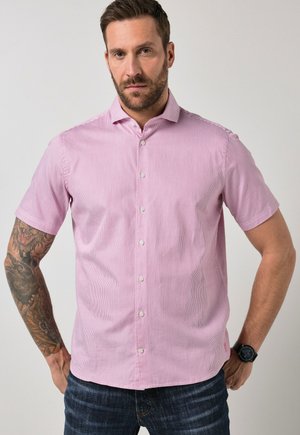 Uomo con barba e tatuaggi che indossa una camicia rosa a maniche corte con bottoni e jeans scuri, in piedi con le mani sui fianchi davanti a uno sfondo semplice.