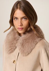 Abrigo beige con suave y esponjoso cuello de pelo marrón y cierre de botón redondo. Textura suave, diseño ajustado. Texto promocional mínimo en la parte superior.