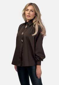 Brązowa koszula zapinana na guziki z wysokim kołnierzem, dużymi srebrnymi guzikami i bufiastymi rękawami; gładka tekstura, w połączeniu z czarnymi skórzanymi spodniami.