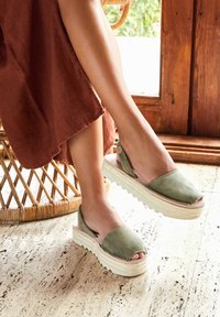 Sandalias de esparto verde de ante con diseño de punta al aire, plataforma elevada y entresuela envuelta en yute, exhibidas sobre un suelo texturizado.
