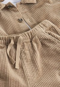 Tan corduroy outfit met een colbert met twee knopen en bijpassende broek met elastische tailleband en trekkoord. Verticale ribbelstructuur.
