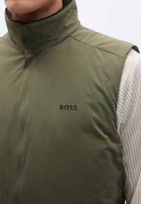 Bez rękawów kamizelka w oliwkowej zieleni z wysokim kołnierzem, gładką fakturą, z czarnym logo "BOSS" na piersi i pasiastym materiałem pod spodem.