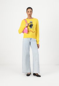 Pull jaune avec un dessin de dessin animé sur le devant, associé à un jean large bleu clair et des chaussures plates noires ; tenant un sac à main rose.