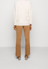 Osoba ubrana w białą, teksturowaną bluzkę z długim rękawem, brązowe spodnie w prążki i sandały na grubym obcasie w zebrę, stoi tyłem na jednolitym tle.