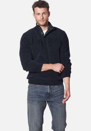 Marineblauer Strickpullover mit gerippten Bündchen, einem Stehkragen und zwei Druckknöpfen am Ausschnitt. Über einer hellblauen Jeans getragen.