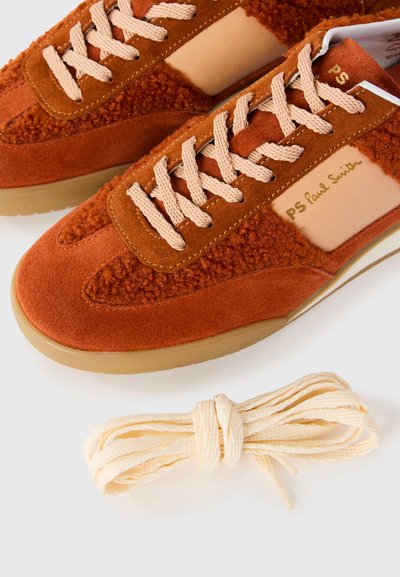 Sneakers in suede arancione ruggine con lacci beige e un set di riserva di lacci color crema su uno sfondo bianco.