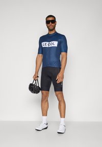 Maillot de cyclisme bleu marine avec le logo blanc "LE COL", short noir, chaussures blanches, tenant un casque noir. Manches courtes, design ajusté, matériau évacuant l'humidité.