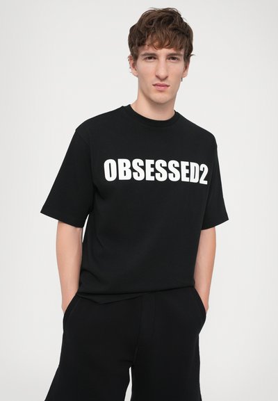 T-shirt en coton noir avec "OBSESSED2" en lettres blanches en gras sur le devant. Manches courtes, coupe décontractée, texture lisse.