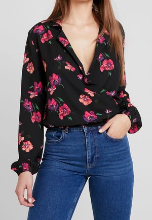 Kobieta w czarnej bluzce z guzikami w czerwono-różowy kwiatowy wzór, w połączeniu z niebieskimi, wysokimi dżinsami, z jedną ręką w kieszeni.