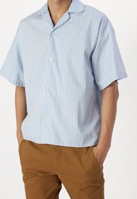 Jasnoniebieska koszula z krótkim rękawem w pionowe białe paski, z kołnierzykiem i guzikami z przodu. W zestawieniu z brązowymi spodniami. Gładka tekstura tkaniny.