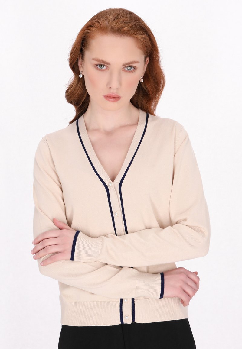 Beige Cardigan mit marineblauem Besatz, V-Ausschnitt und Knopfleiste. Hat lange Ärmel, gerippte Bündchen und eine glatte Textur, getragen über schwarzen Hosen.