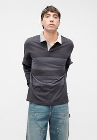 Νεαρός άνδρας που φοράει γκρι σκούρο ριγέ ράγκμπι πουκάμισο με μπεζ γιακά, μπλε τζιν με μεγάλες μπροστινές τσέπες και μαύρο καπέλο φορεμένο ανάποδα, όρθιος.