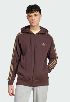 Sudadera con cremallera Burgo marrón con rayas doradas a lo largo de las mangas, capucha con cordón, textura suave y logo bordado en el pecho. Combinada con pantalones grises.