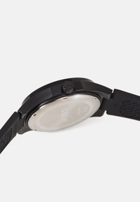 Black analog watch with a matte finish. Features a round case, textured strap, and a silver metal case back labeled "HUGO."