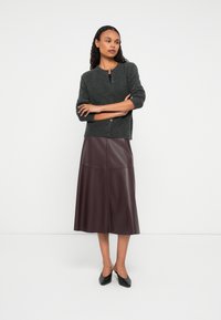 Donna in piedi con le braccia incrociate, indossa un cardigan grigio scuro, una gonna midi in pelle bordeaux e scarpe nere con tacco basso, su sfondo bianco.