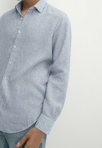 Man in een lichtblauw en wit verticaal gestreept overhemd met lange mouwen en omgeslagen mouwen, gecombineerd met lichtblauwe jeans.