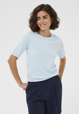 Rövid barna hajú nő mosolyog, világoskék rövid ujjú felsőt és sötétkék magas derekú nadrágot visel, egyszerű háttér előtt áll.