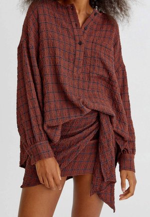 Kobieta ubrana w długorękawową bluzkę w kratę w kolorach rdzawego i granatowego oraz dopasowaną mini spódniczkę z zakładką na tle jednolitego tła.