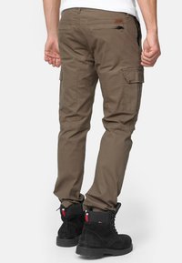 Brązowe spodnie cargo wykonane z wytrzymałego materiału, z bocznymi kieszeniami, slim fit i w połączeniu z czarnymi butami. Tekstura wydaje się gładka i solidna.