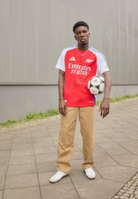Maillot de soccer Adidas rouge et blanc avec le logo "Emirates Fly Better". Pantalon large beige et baskets blanches. Tient un ballon de soccer noir et blanc.