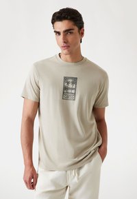 Lichtbeige t-shirt gemaakt van zachte katoen, met een zwart grafisch ontwerp van palmbomen in een rechthoekig kader op de borst.