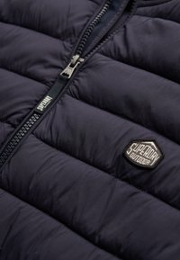 Gros plan sur une veste matelassée foncée avec une fermeture éclair frontale et un patch logo « Superdry Outdoor » sur la poitrine.